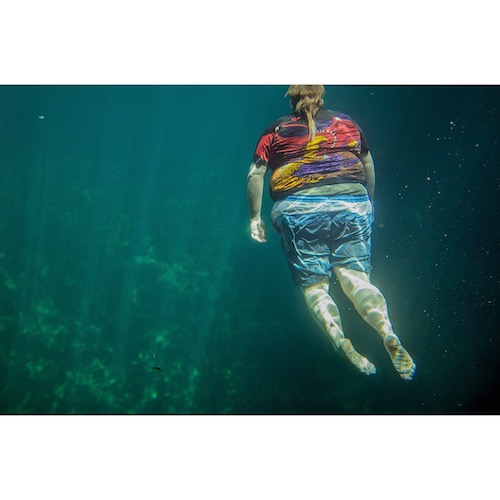 Foto av queer konstnär Lo River Lööf. En person flyter under vatten med shorts och T-shirt, med ryggen mot oss och ljus strilandes genom vattnet.