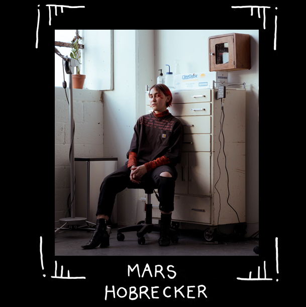 Fotoporträtt av queer tatuerare Mars Hobrecker som sitter på kontorsstol framför ett skåp, nedanför ett högt fönster. Mars sitter avslappnat och tittar in i kameran, bredbent med klackskor och trasiga byxor. Plastflaskor och powersupply syns i inredningen, det ser ut att vara arbetsplats.