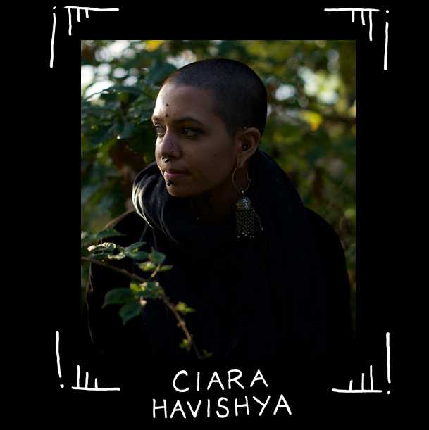 Fotoporträtt av queer tatuerare Ciara Havishya. Ciara är allvarlig och sneglar åt sidan, står framför grönt lummigt, kanske rosensnår, med svart munkjacka, rakat huvud och septum piercing.