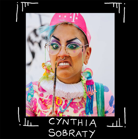 Fotoporträtt av queer tatuerare Cynthia Sobraty. Me lila ögonskugga och kantigt målade ögonbryn, rosa cykelkeps och färgglad clownliknande dräkt. Tittar på pensel med arg min.