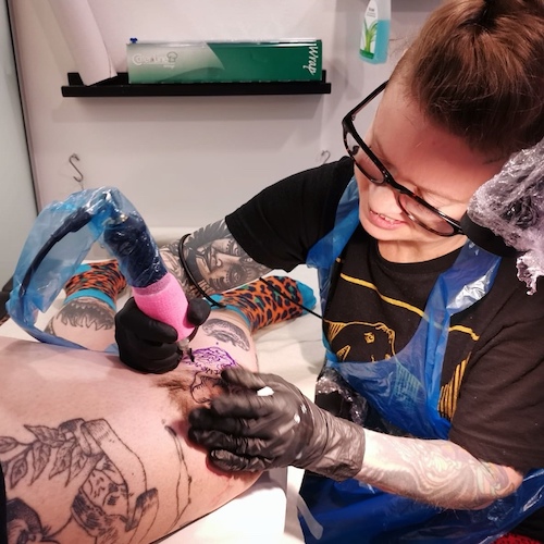 Queer tatuerare Sara tatuerar på sidan av ett knä, en fladdermus. Sara har plastförkläde och stora glasögon på sig.