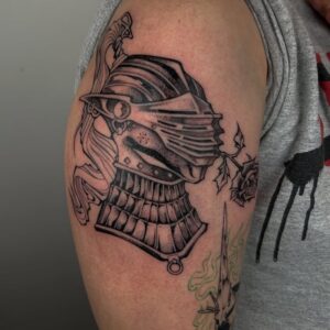 En riddarhjälm med ros i munnen är tatuerad över en överarm. Tatueringen är gjord i old school stil av tatuerare Milena Huhta.