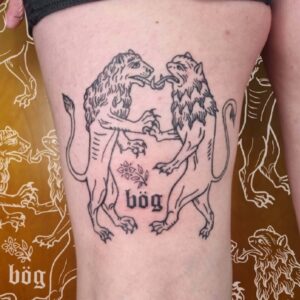 Två lejon tatuerade i fine line medeltid stil, likt gravyr. Tatuering över lår. Lejonen står upp och snirklar ihop sina tungor. Mellan dem står texten Bög tatuerad. Av queer tatuerare Petrix.