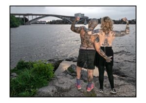 Two persons showing their tattooed naked backs, with bridges and water as a background in Stockholm.